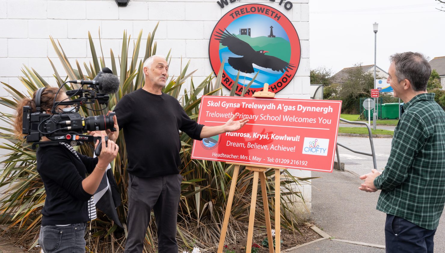 A Man stands in front of a Bilingual Cornish & English School Sign while being interviewed, a television camera is being held up to his side