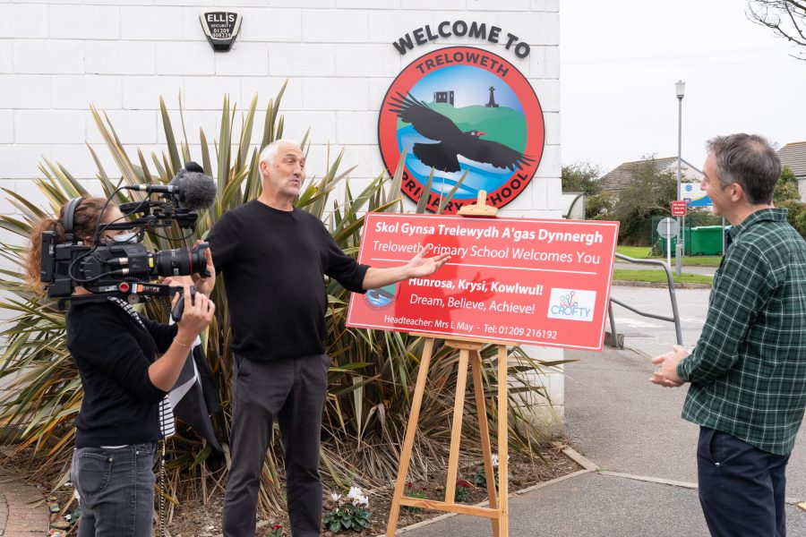 A Man stands in front of a Bilingual Cornish & English School Sign while being interviewed, a television camera is being held up to his side