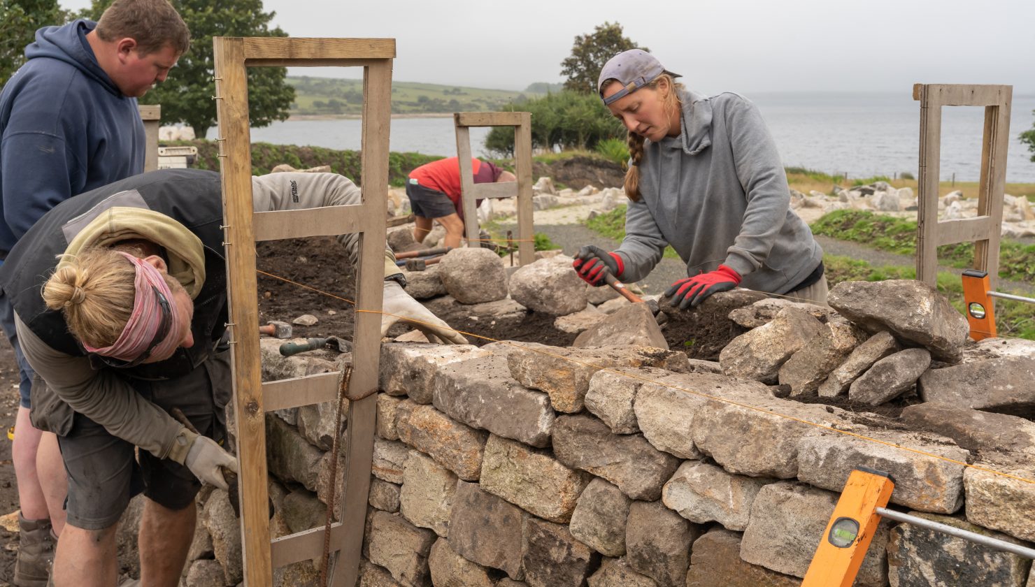 A group of people wearing work gloves are leaning over a stone hedge construction propped up with wooden former